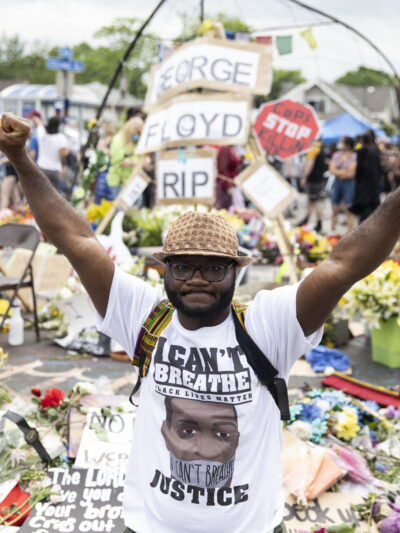 Man wearing "I Can't Breathe" tshirt stands in front of George Floyd memorial with fists raised.