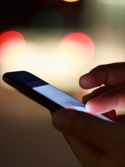 A pair of hands holding a cell phone at night with street lights in the background.