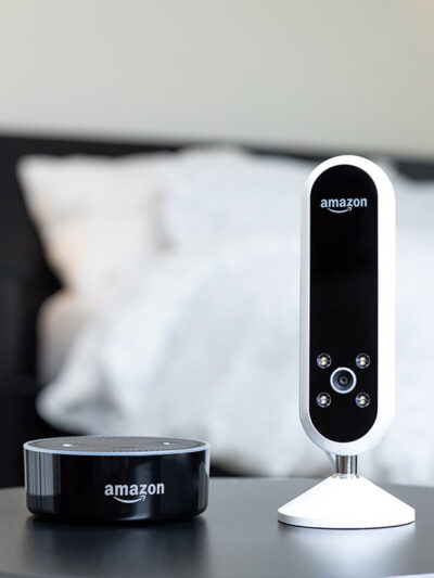 Amazon camera and Amazon Alexa speaker next to each other on a bedside table.