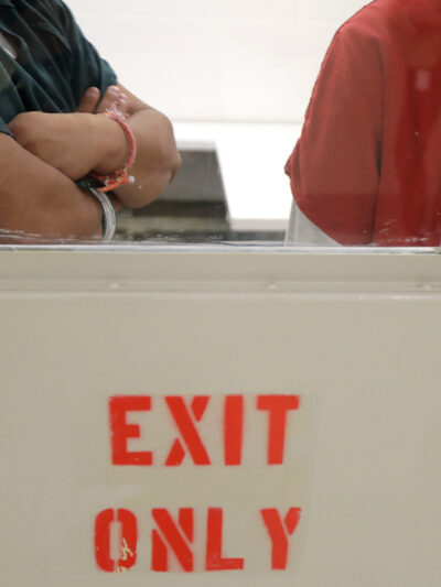 Two detainees seen waiting through a window behind closed doors.