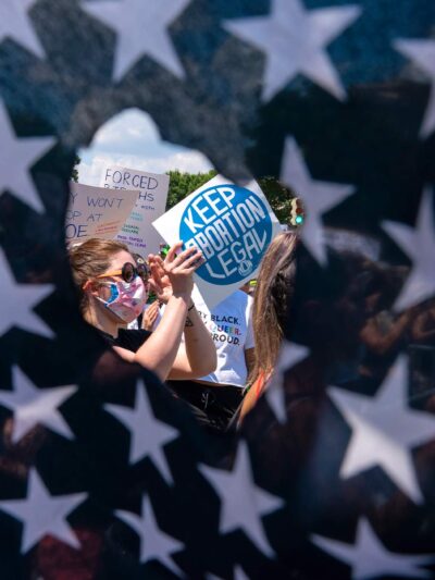 Abortion rights activists are seen through a hole in an American flag as they protest outside the Supreme Court in Washington, Saturday, June 25, 2022.