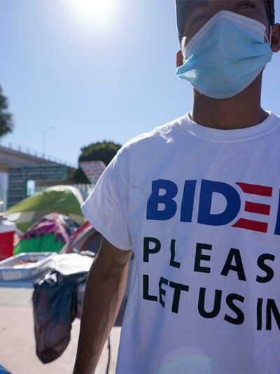 A man seeking asylum in the United States wears a shirt that reads, "BIDEN PLEASE LET US IN!," as he stands among tents that line an entrance to the border crossing in Tijuana, Mexico.