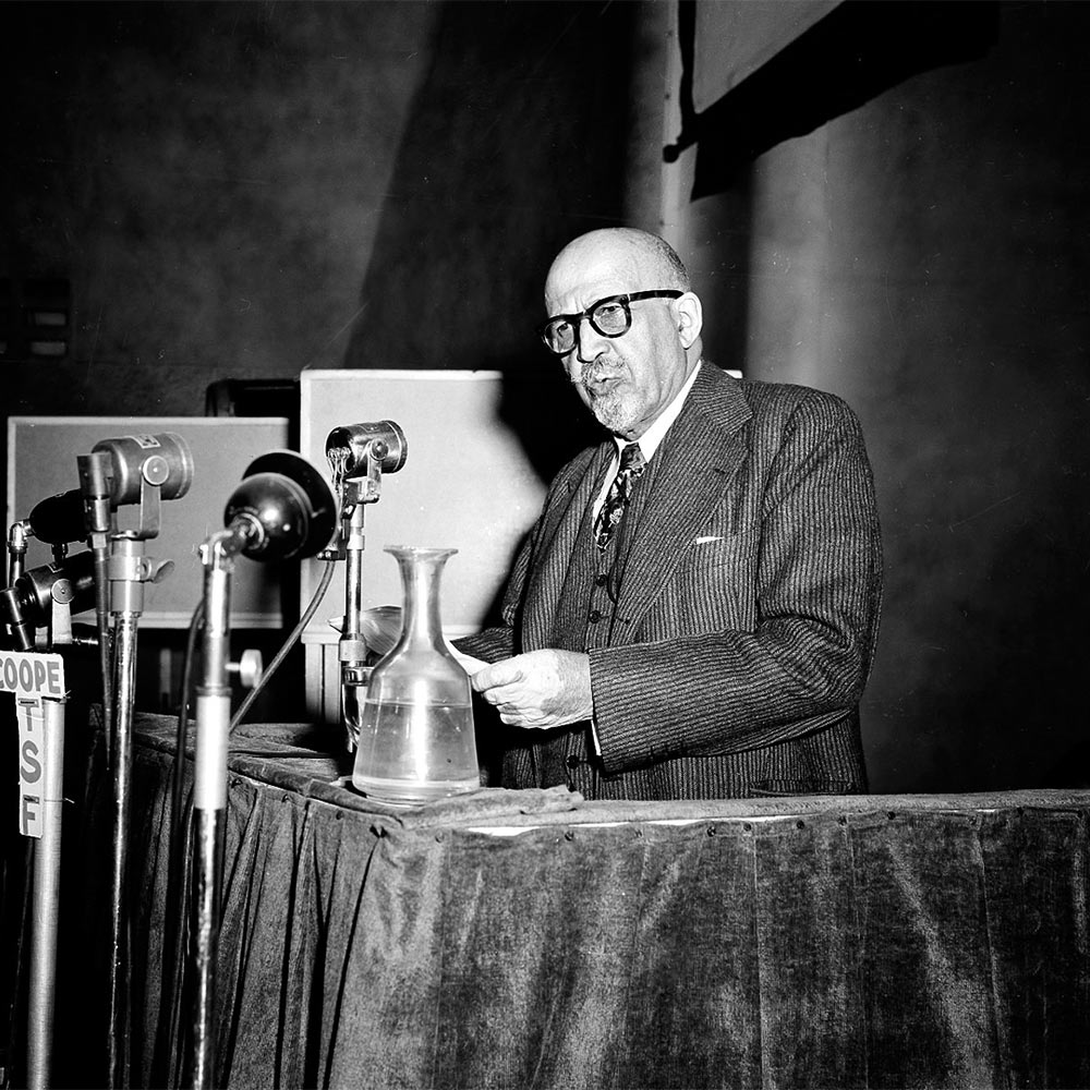 A black and white photo of W.E.B. DuBois standing at a podium in front of multiple microphones.