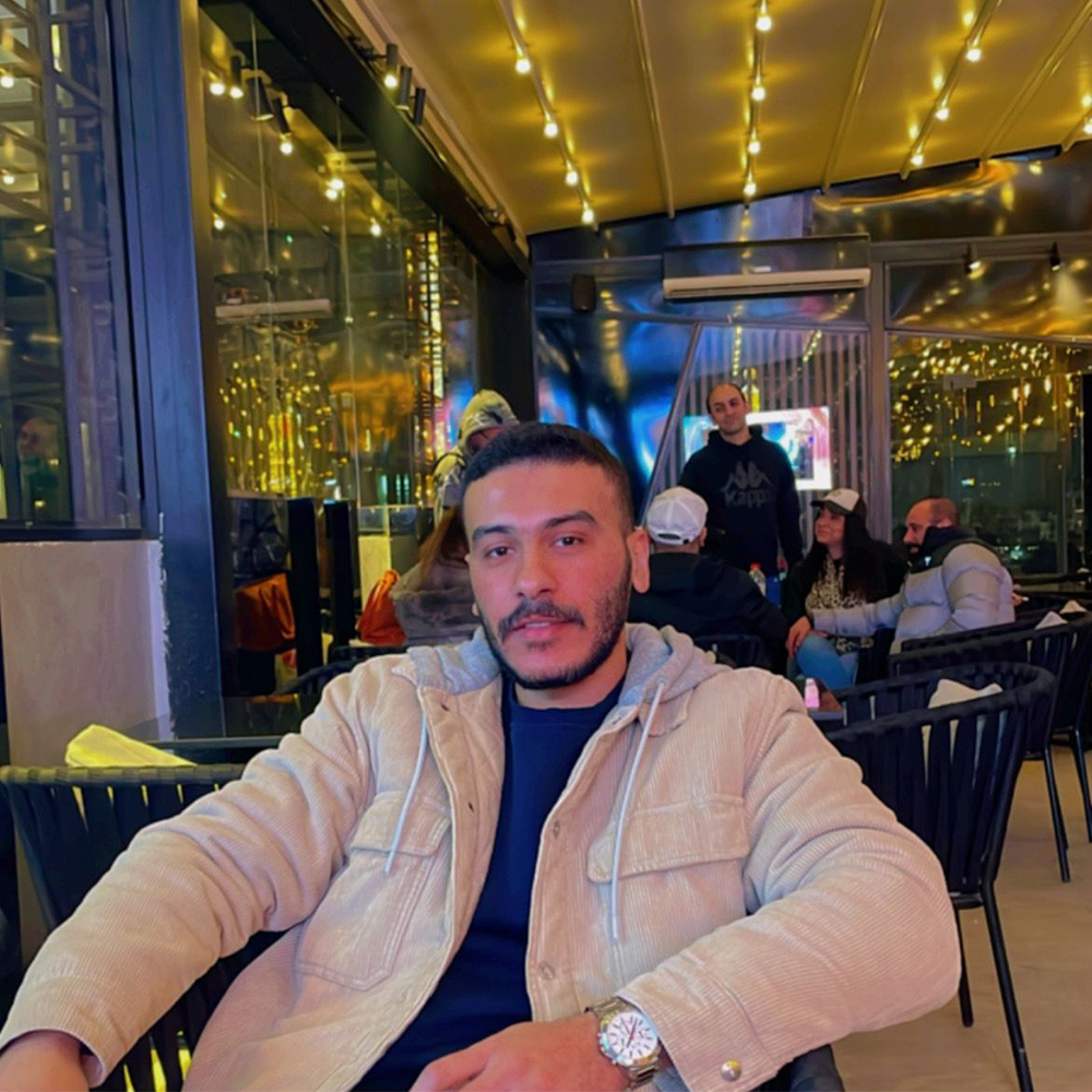Moataz Abdelazim sits at a bistro table in a restaurant with warm lighting. He is wearing an off-white corduroy jacket with a hood, layered over a blue t-shirt with a gold wrist watch. He has short brown hair with a fade along the perimeter.