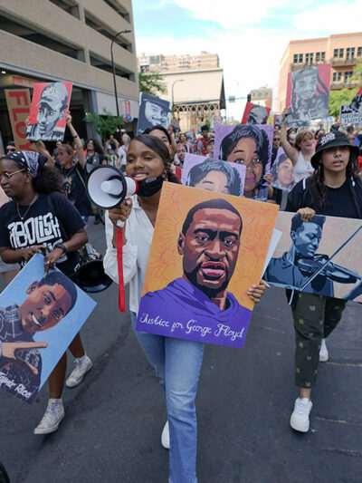 Protestors walking down the street, with one holding a microphone and a sign with art of George Floyd's face.