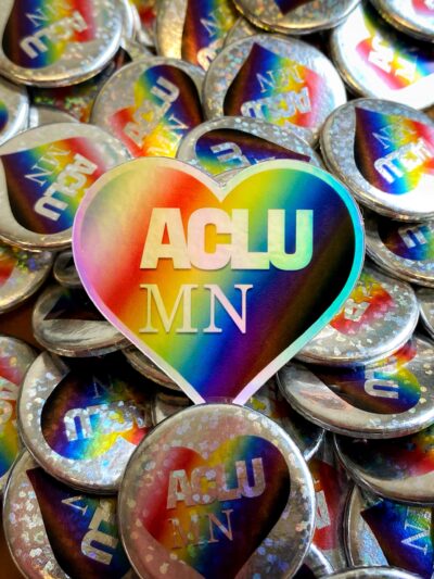 Heart-shaped sticker with rainbow pattern and white "ACLU MN" text sitting in the middle of a pile of silver pins with the same design
