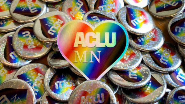 Heart-shaped sticker with rainbow pattern and white "ACLU MN" text sitting in the middle of a pile of silver pins with the same design