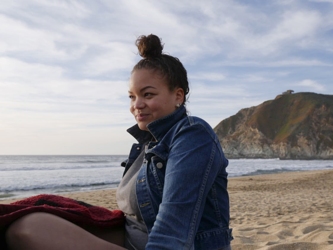 Photo of Brittany wearing a jean jacket and shorts and sitting on a sandy beach.