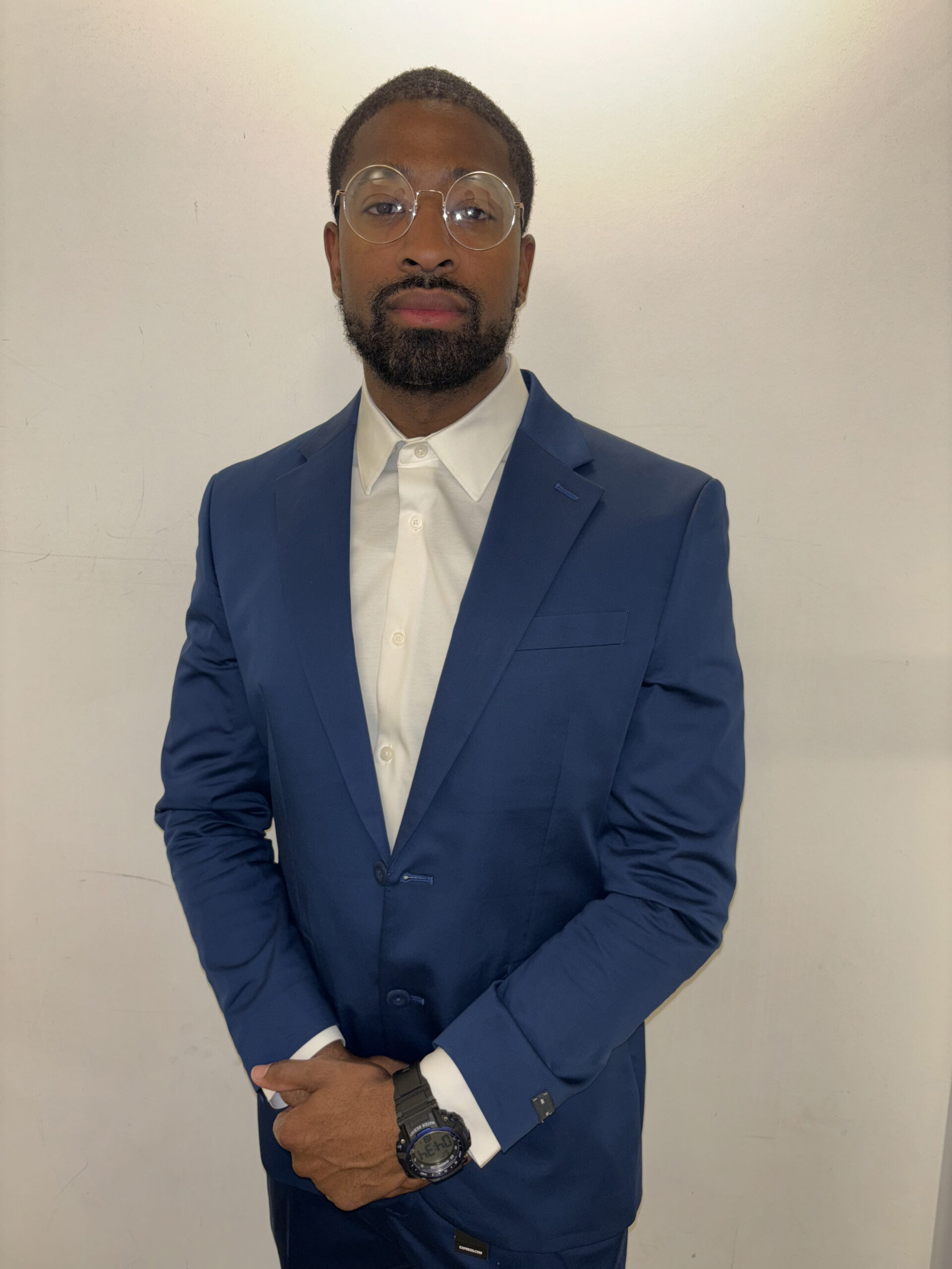 Photo of Calvin in a navy blue suit and a white shirt against a cream wall.