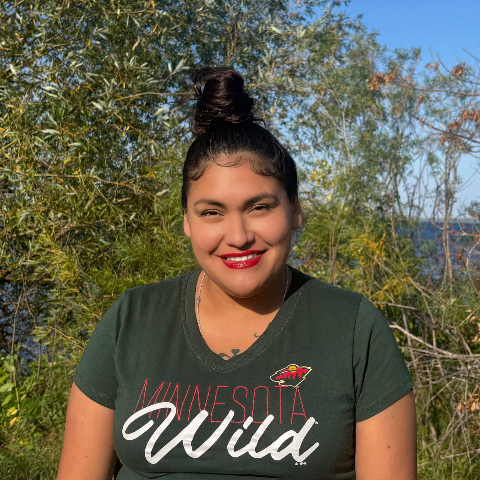 Headshot of Winona wearing a dark t-shirt and bright red lipstick sitting in front of trees.