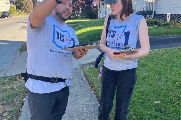 Photo of Julio on left pointing and talking to a woman on the right. Both are wearing gray t-shirts that say "yes on issue 1" with the shape of the state of Ohio behind the text.