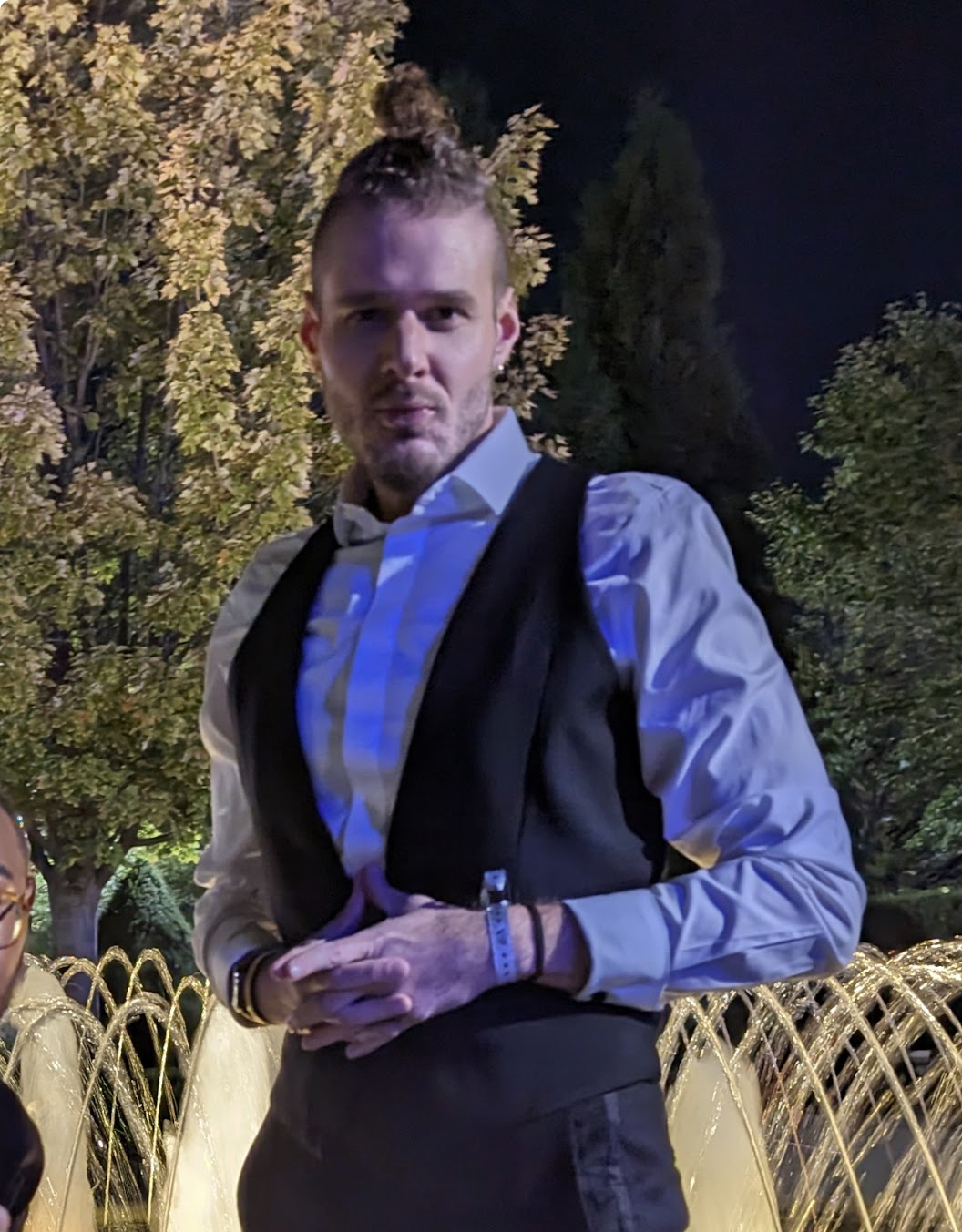 Photo of Oleg wearing a black vest and white button down with trees in the background.