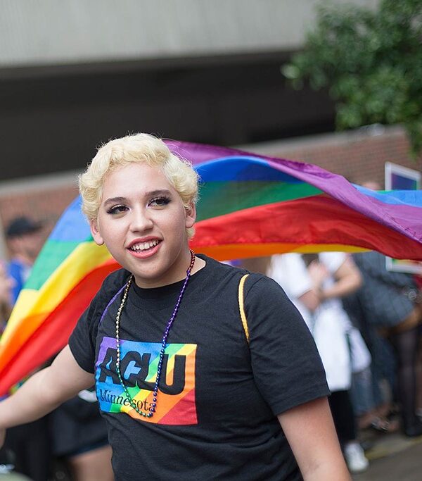 Photo of person wearing black t-shirt that says "ACLU Minnesota" and carrying a Pride flag that flows behind them.