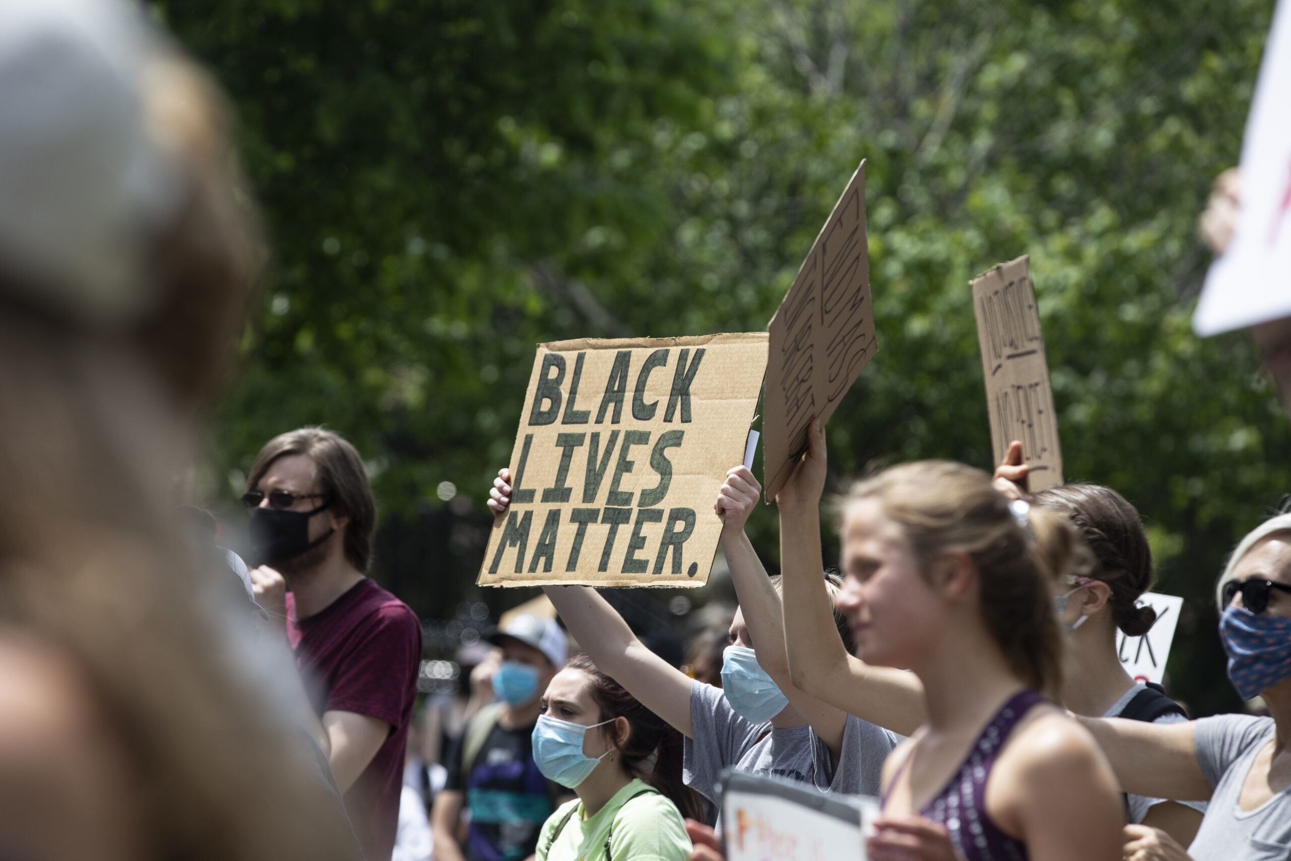Photo of people protesting and holding homemade signs. The sign in the middle says, "Black Lives Matter."