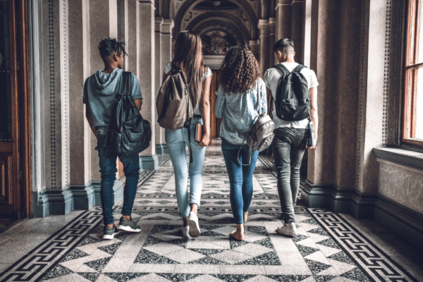 Photo of four college students wearing backpacks walking down a hallway with marble floor.