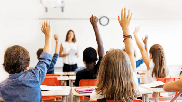 Photo of a classroom with students raising their hands and facing the teacher