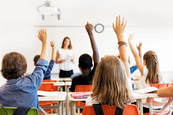 Photo of a classroom with students raising their hands and facing the teacher
