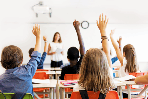 Photo from the back of a classroom where students are sitting in desks and raising their hands.