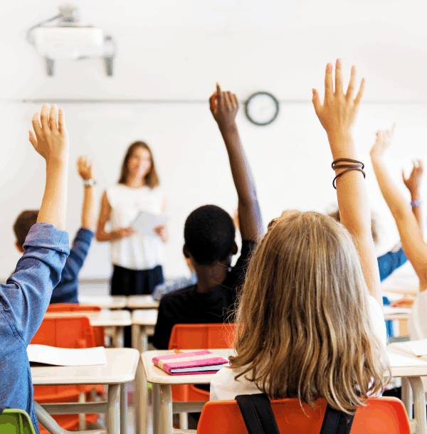 Photo from the back of a classroom where students are sitting in desks and raising their hands.
