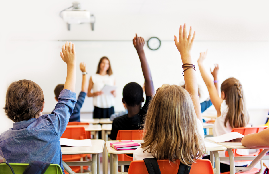 Photo from the back of a classroom where students are sitting in desks and raising their hands.