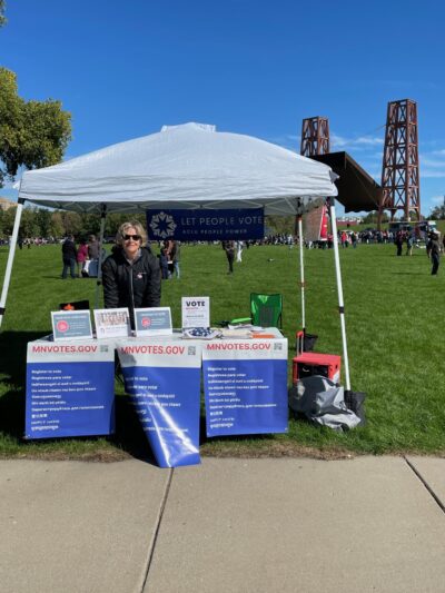Photo of an individual with short blond hair, wearing sunglasses, standing under a white tent. In front of the person is a table with signs attached to it. The signs say, "MNvotes.gov."