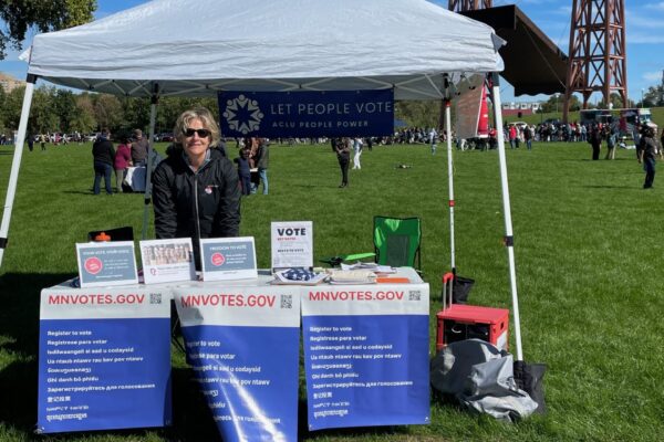 Photo of an individual with short blond hair, wearing sunglasses, standing under a white tent. In front of the person is a table with signs attached to it. The signs say, "MNvotes.gov."