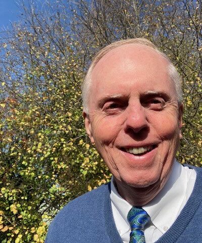 Photo of Bruce wearing a blue sweater and tie in front of a small, green tree.