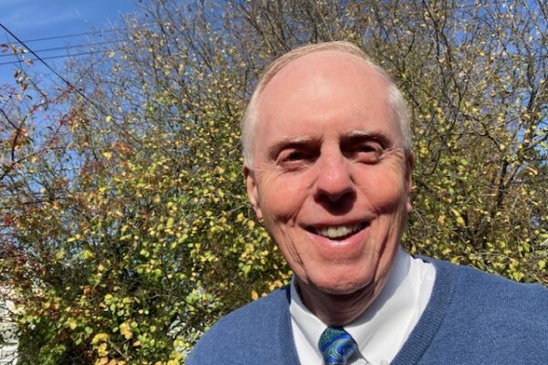 Photo of Bruce wearing a blue sweater and tie in front of a small, green tree.