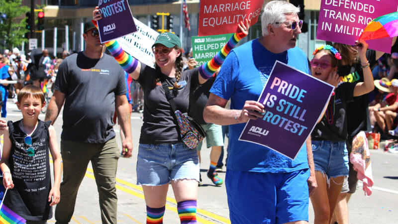 Photo of people marching in a parade. One person has a sign that says, "ACLU Minnesota defends equal education for trans teens."