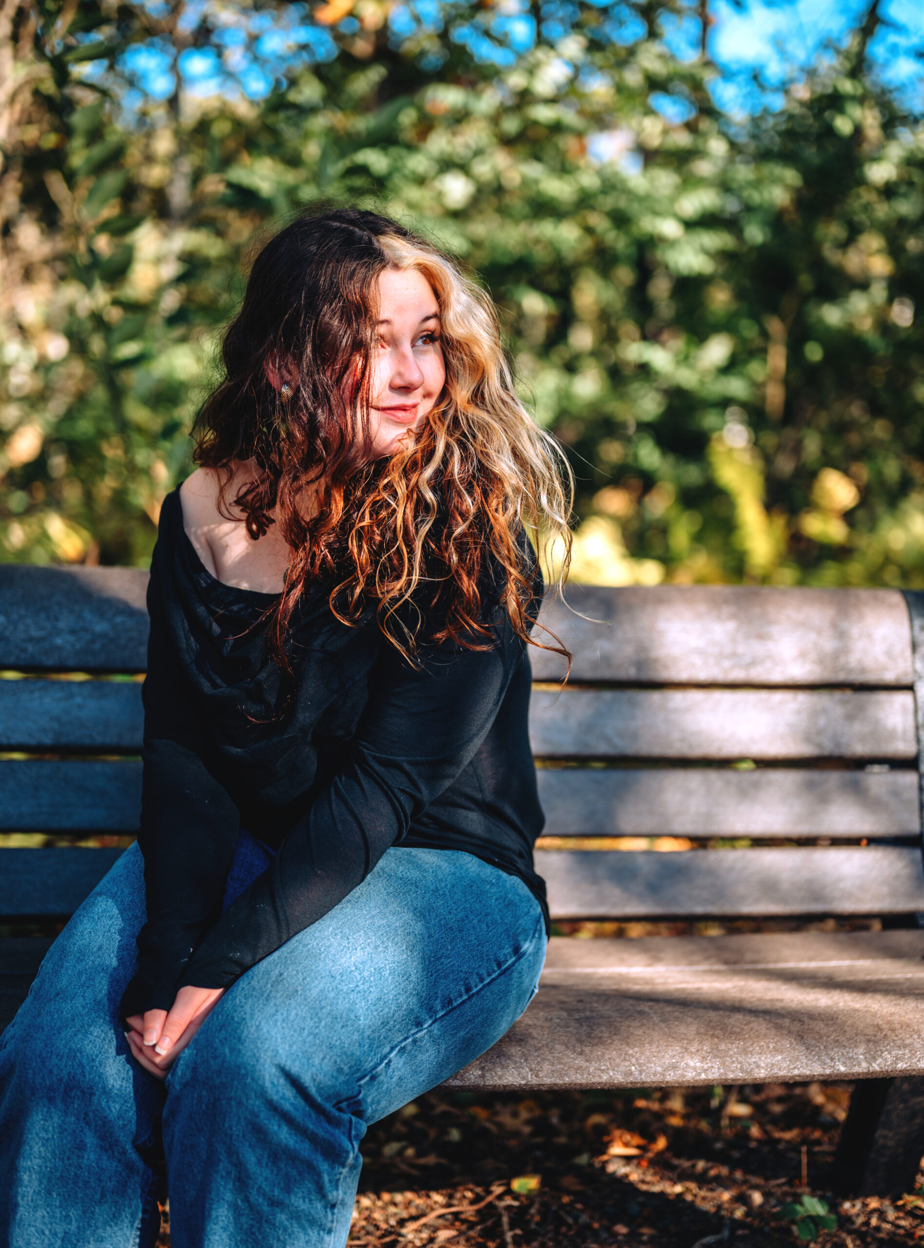 Photo of Rowan sitting on a bench outside wearing a black long sleeve shirt and blue jeans.