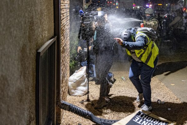 Photo of a journalist wearing a yellow blazer that says "press" and a person carrying a video camera shielding their eyes as an officer sprays them with pepper spray