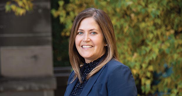 Photo of Catherine Ahlin-Halverson. A white woman with a bob haircut and a navy blue blazer poses in front of a blurred outdoor background and smiles at the camera.