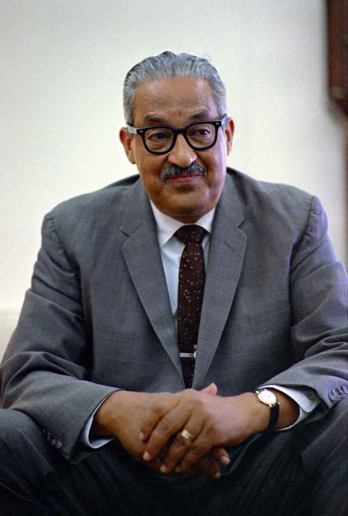 Image of Thurgood Marshall looking off camera with hands crossed. He is wearing a gray suit and a dark patterned tie with a wrist watch on his left wrist.