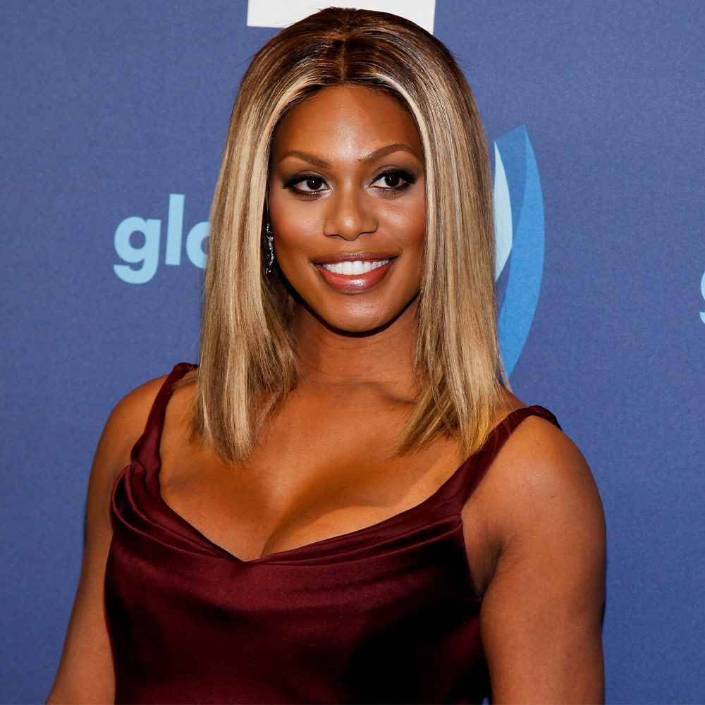 Actress Laverne Cox wearing a burgundy dress stands in front of a blue photo backdrop while smiling and looking off camera.
