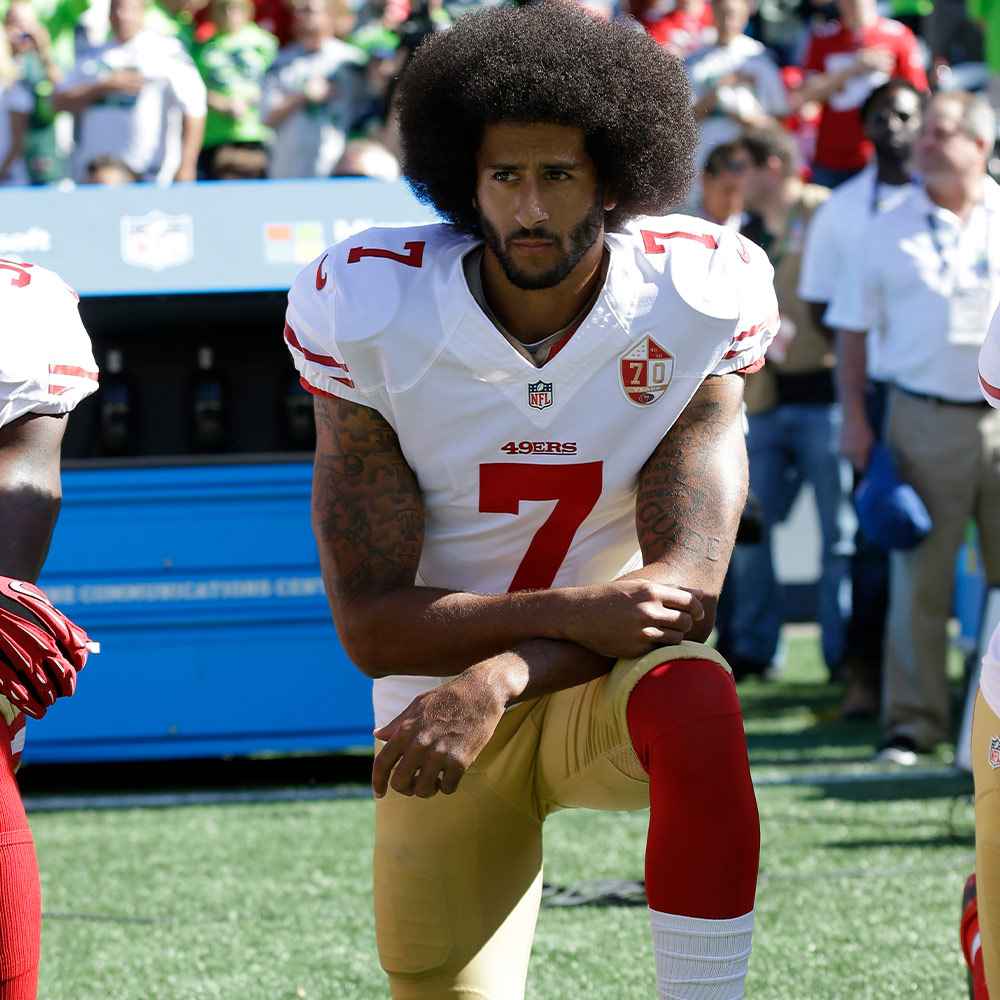 Iconic image of quarterback Colin Kaepernick kneeling at a football game. He is wearing his red, white and gold San Francisco 49ers uniform.