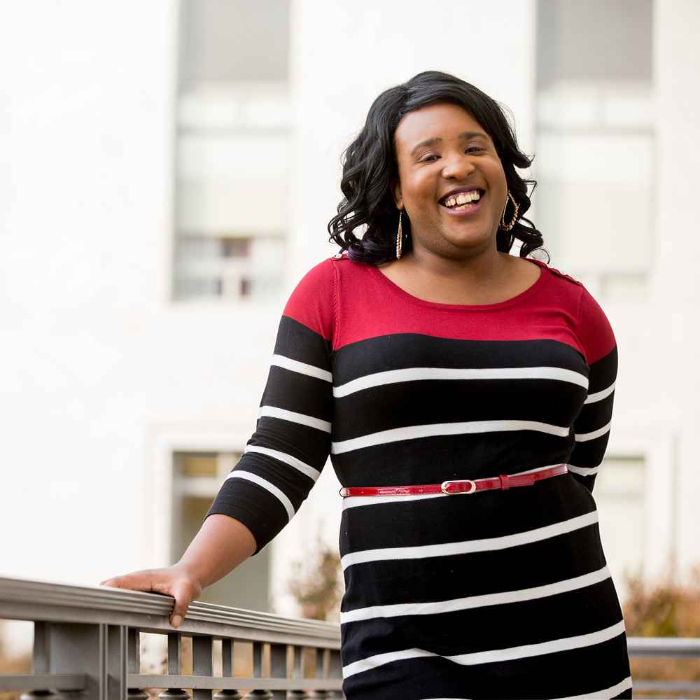 Candid photo of Lala Zannell smiling with one hand leaning against a railing. She is wearing a black dress with horizontal red and white stripes belted at the waist and has her hair in full curls. 