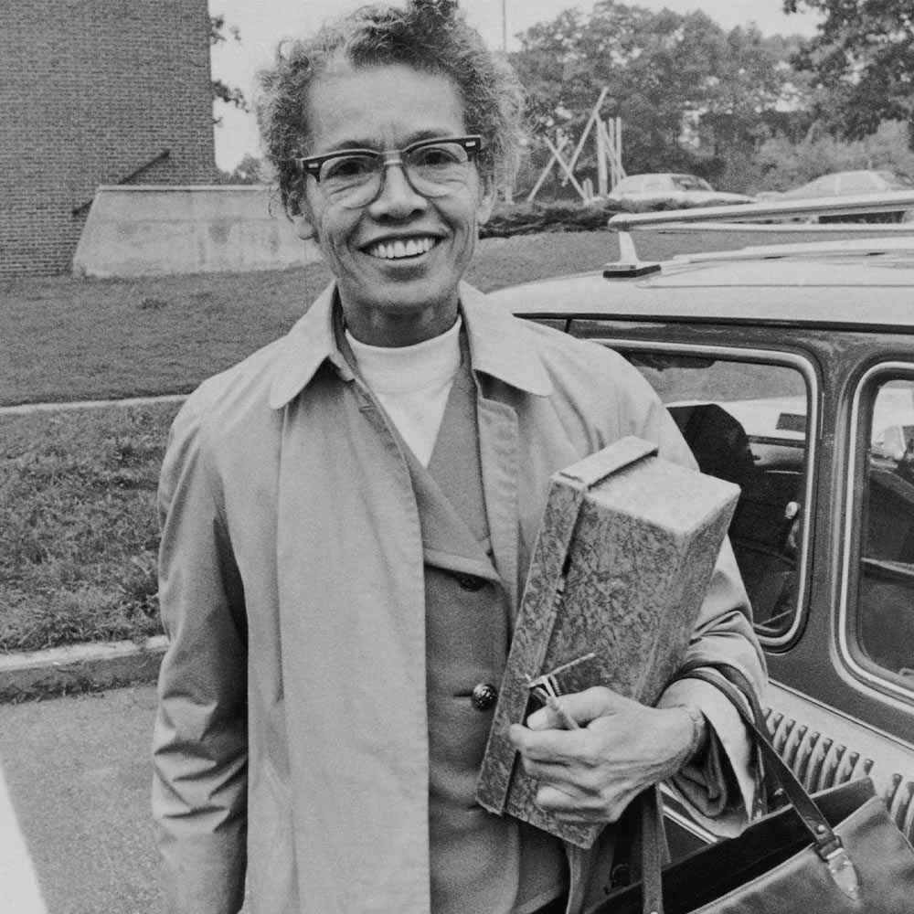 Pauli Murray, wearing an overcoat and blazer, stands in front of a car while holding a box and smiling.
