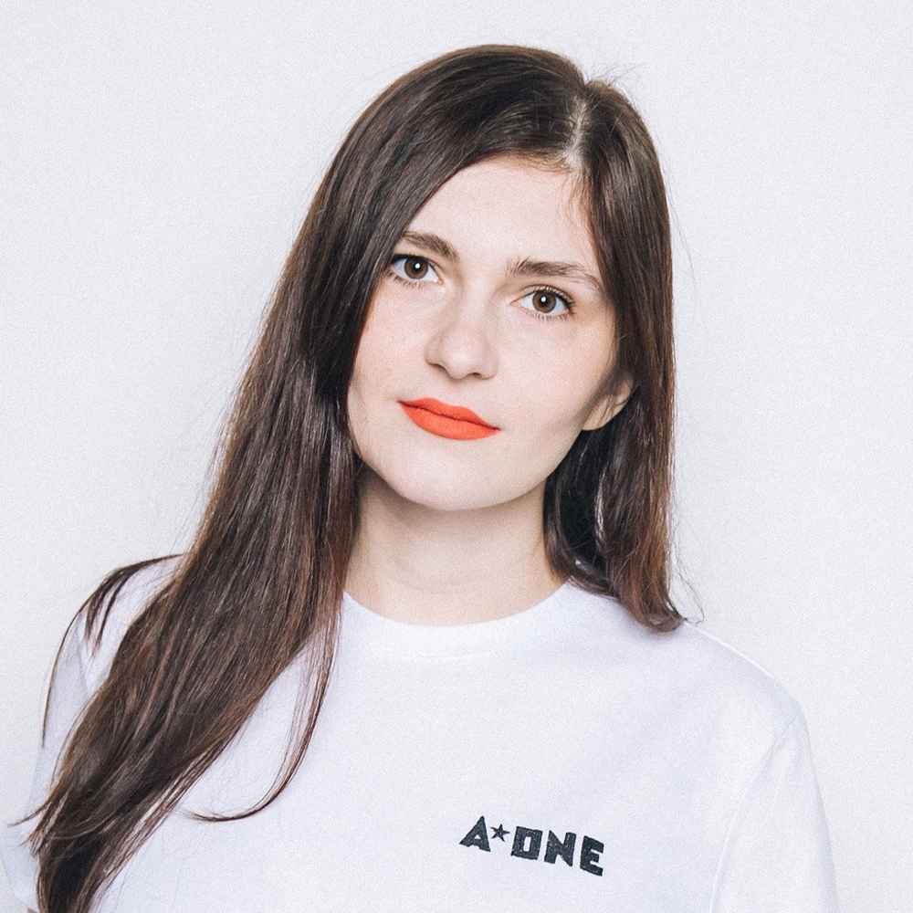 Ekaterina Karslidi wears a white t-shirt that says "A-ONE" in front of a white backdrop. She is a long-haired brunette with brown eyes and is wearing an orange lipstick.