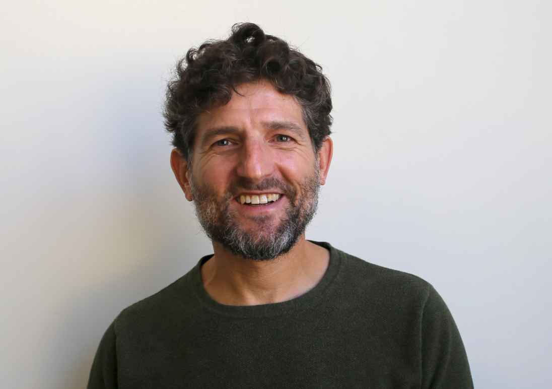 Photo of Ben wearing a dark green sweater standing against a white wall. 