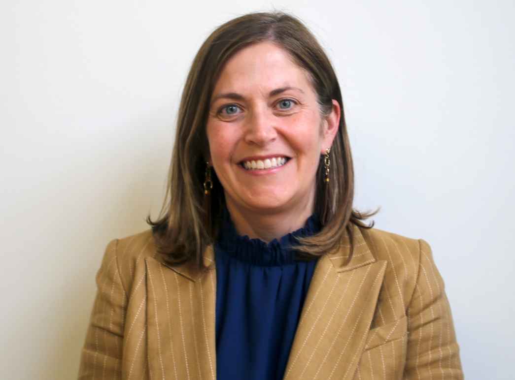 Catherine wearing a dark top and tan jacket against a white wall. 