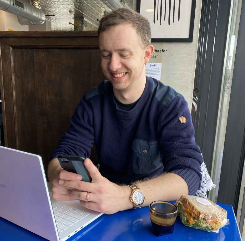 Photo of Paul sitting in a coffee shop with his computer and phone