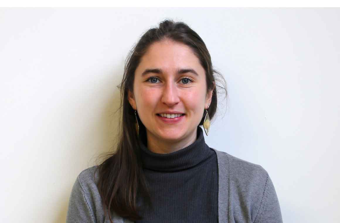 Grace wearing a black shirt with grey cardigan stands against a white wall. 