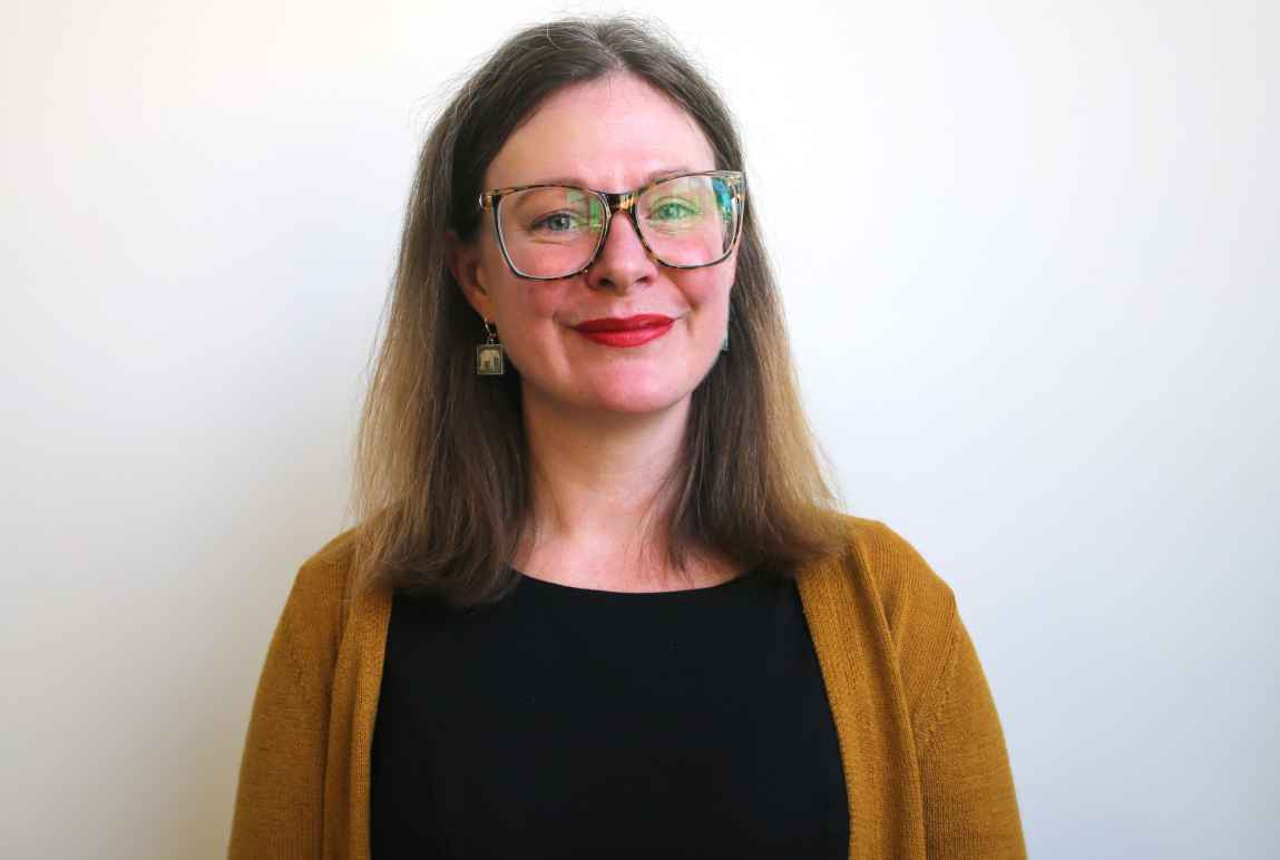 Hailey standing against a white wall wearing a black top and mustard yellow cardigan 