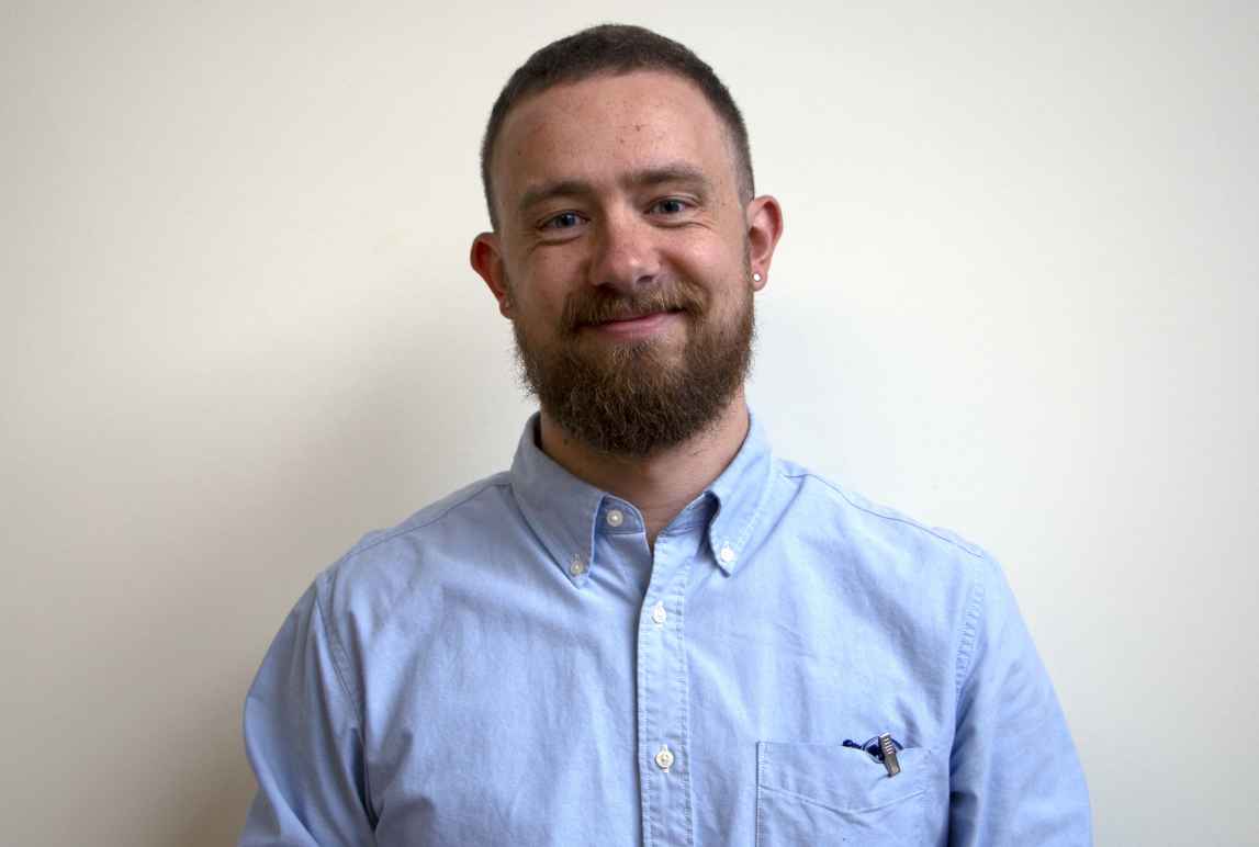 Headshot of Paul standing against a white wall wearing a light blue button-up shirt