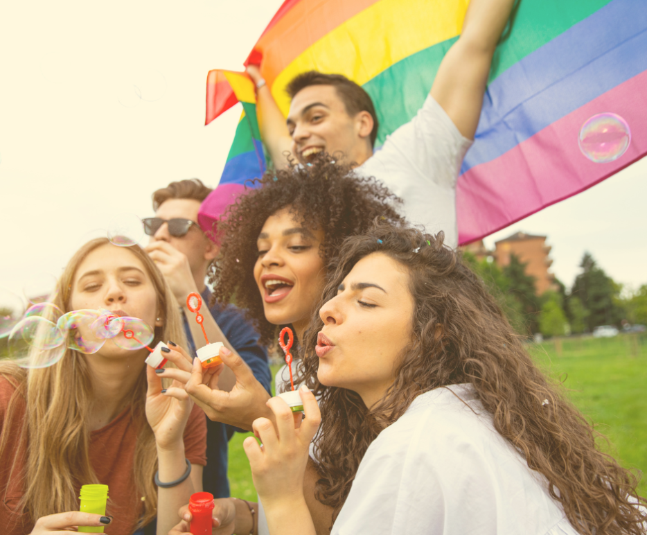 Photo of people sitting on the ground and blowing bubbles and one person holding a Pride flag above all of them.