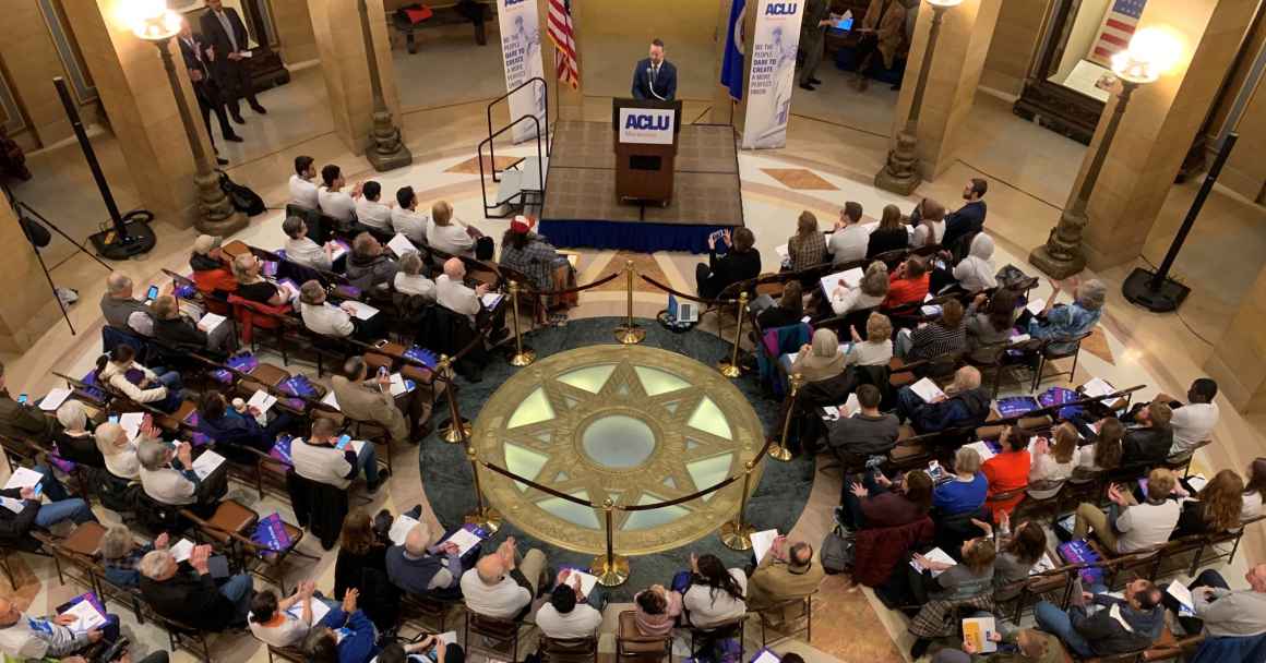 Photo of people sitting in Minnesota Capitol Rotunda listening to someone speak from a small stage.
