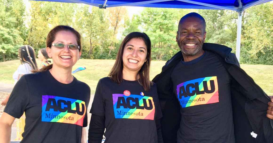 Photo of three people standing next to each other and smiling at the camera. Each person is wearing a dark grey t-shirt with a large, rainbow ACLU of Minnesota logo in the center.