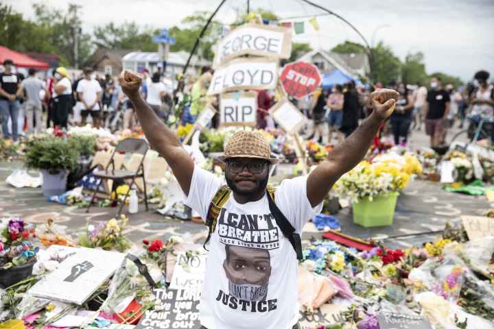Man wearing "I Can't Breathe" tshirt stands in front of George Floyd memorial with fists raised.