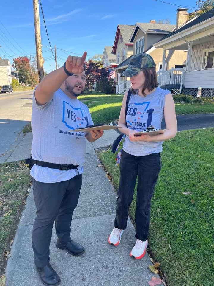 Photo of Julio on left pointing and talking to a woman on the right. Both are wearing gray t-shirts that say "yes on issue 1" with the shape of the state of Ohio behind the text.