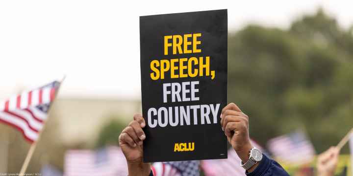 A demonstrator (with only their hands visible) holds up a sign that reads " FREE SPEECH, FREE COUNTRY - ACLU".